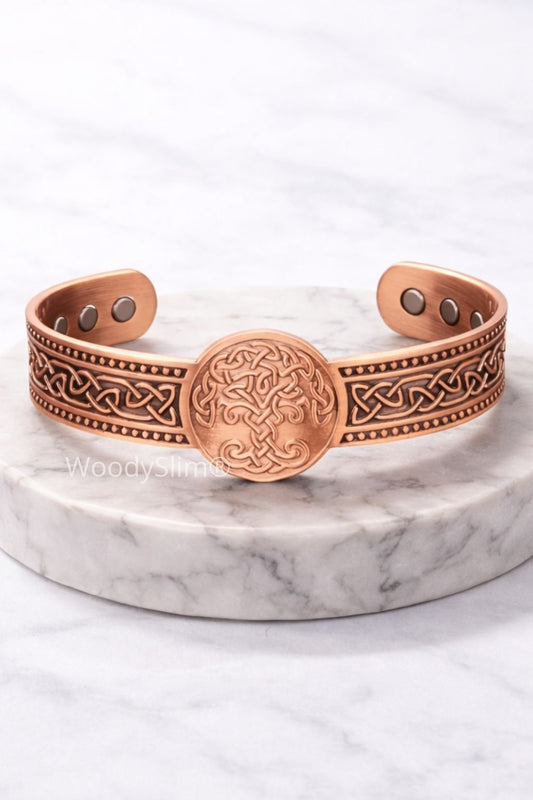 Bracelet en cuivre gravé de motifs celtiques et d’un arbre de vie, posé sur un socle rond en marbre blanc. Fond clair, ambiance minimaliste et élégante, fidèle à l’univers WoodySlim®.