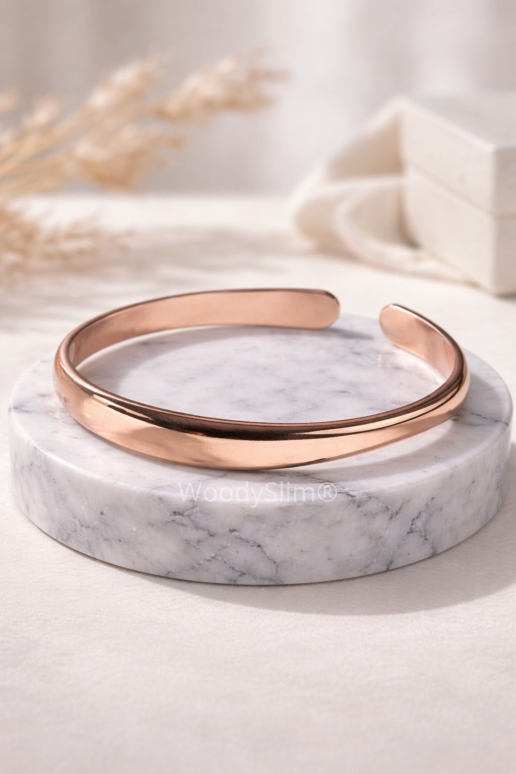Bracelet en cuivre finition polie présenté seul sur un socle en marbre clair, design ouvert et épuré, bijou WoodySlim.