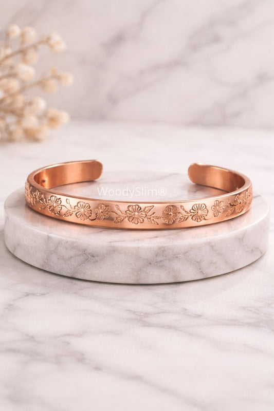 Bracelet en cuivre gravé à motifs floraux, finition cuivrée brillante, posé sur un socle en marbre clair, bijou bien-être élégant WoodySlim