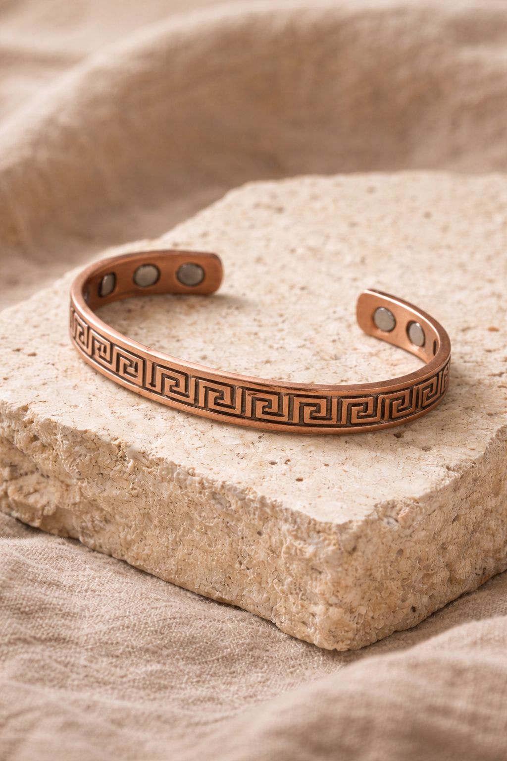 Bracelet en cuivre rigide ouvert avec motif grec gravé et pastilles intérieures visibles, présenté sur un support en pierre claire, bijou WoodySlim.