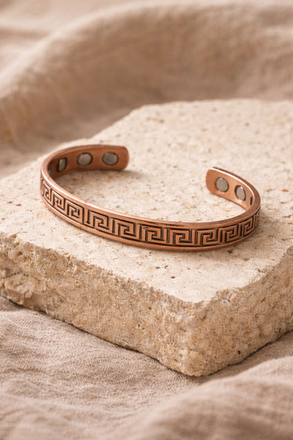 Bracelet en cuivre rigide ouvert avec motif grec gravé et pastilles intérieures visibles, présenté sur un support en pierre claire, bijou WoodySlim.